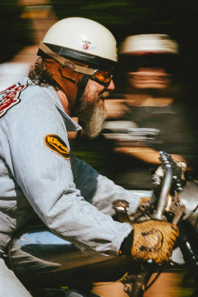Man with a beard wearing a white helmet, sunglasses, and a racing jacket riding a motorcycle, with another rider blurred in the background.