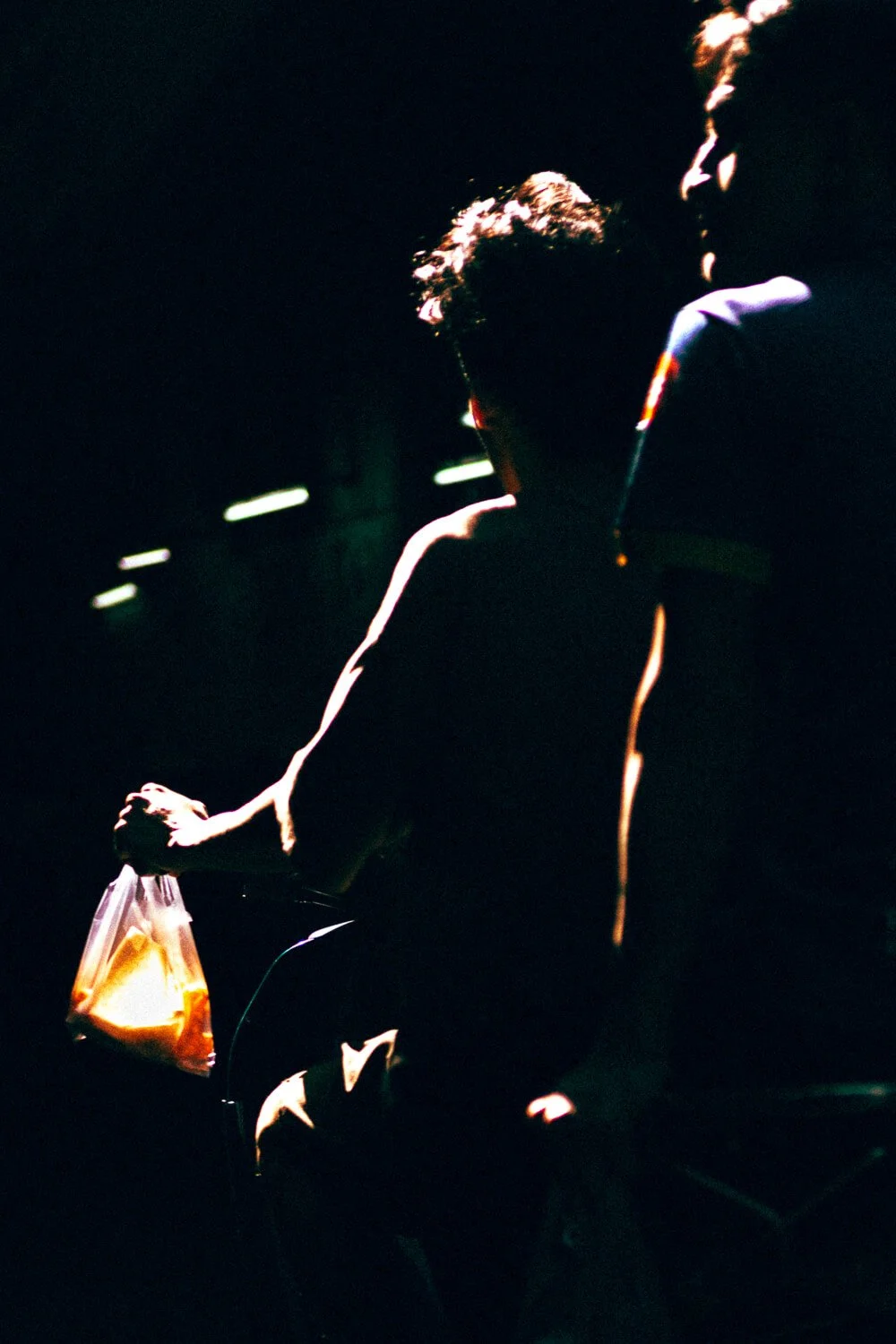 Deux personnes dans une obscurité, l'une portant un sac en plastique, la lumière mettant en valeur leurs silhouettes.