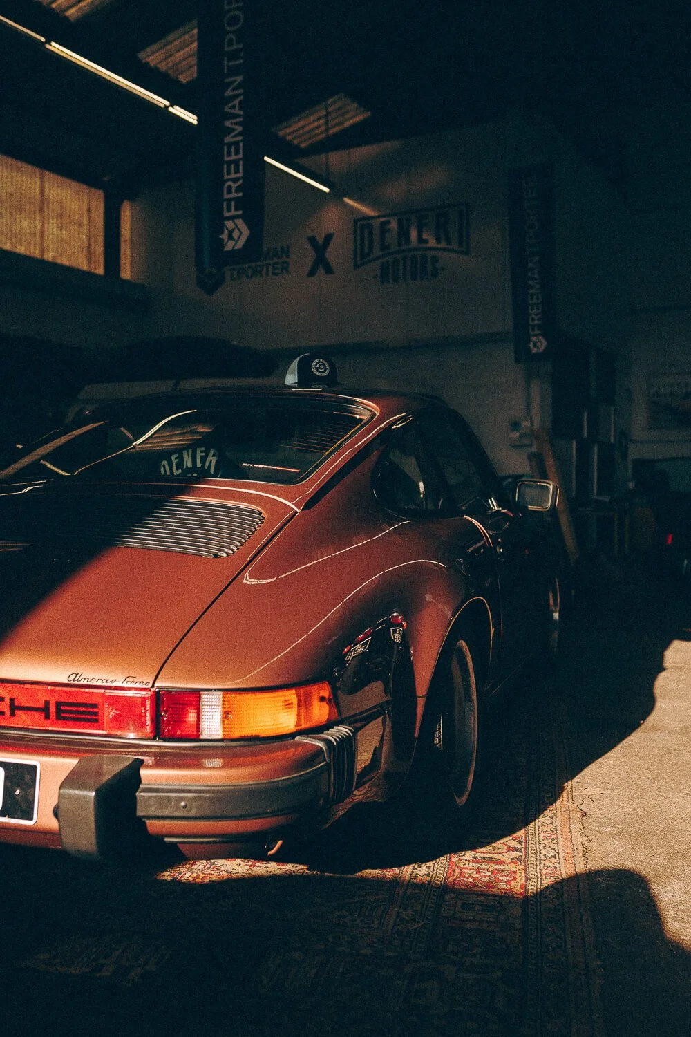 A brown vintage Porsche car parked indoors with a shadow cast over it, banners hanging from the ceiling reading 'FREEHMAN-TPORTER' and 'DEMERI MOTORS'.