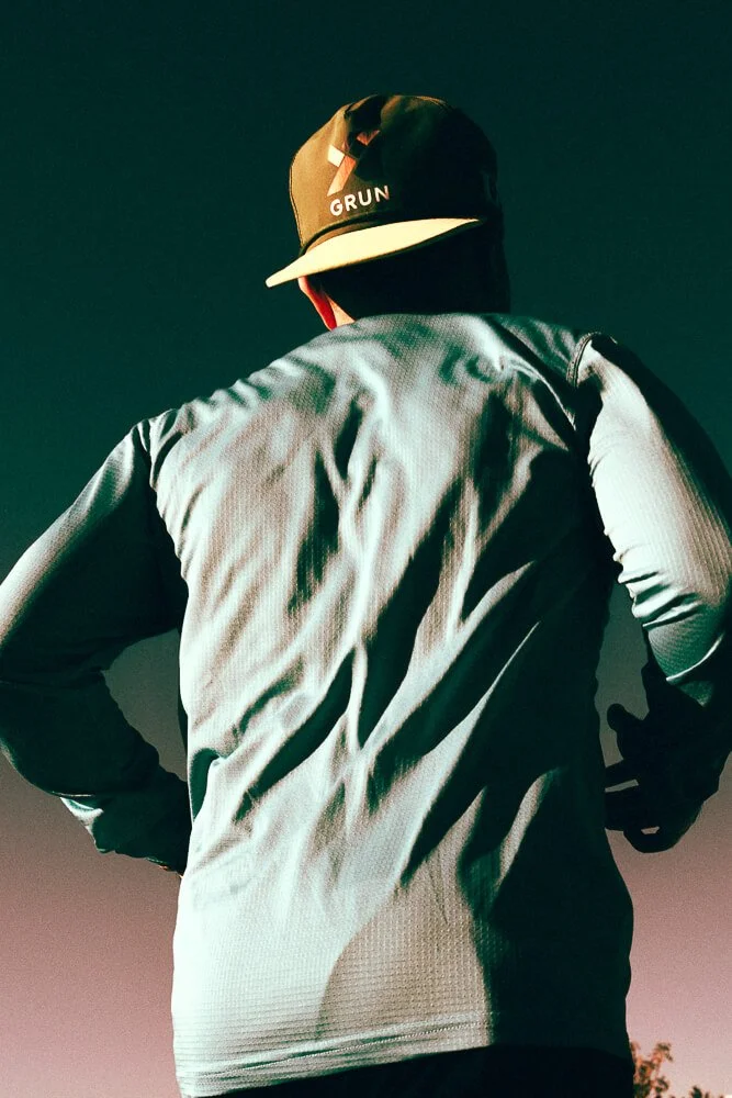 Back view of a person wearing a gray athletic jacket and a black cap with a logo, standing outdoors during sunset or dusk.