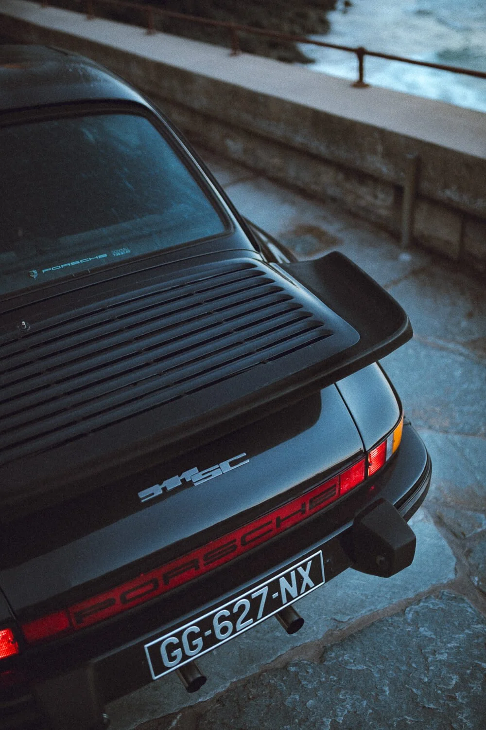 The rear of a classic black Porsche 911 with a rear spoiler, red and white Porsche logo, and French license plate, parked on a stone pavement near a railing by water.