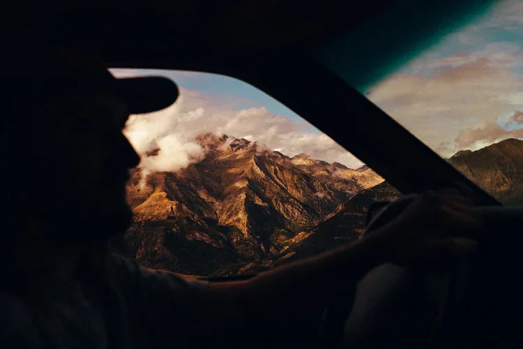 Silhouette d'une personne au volant d'une voiture, vue à travers le pare-brise vers un paysage montagneux au coucher du soleil.