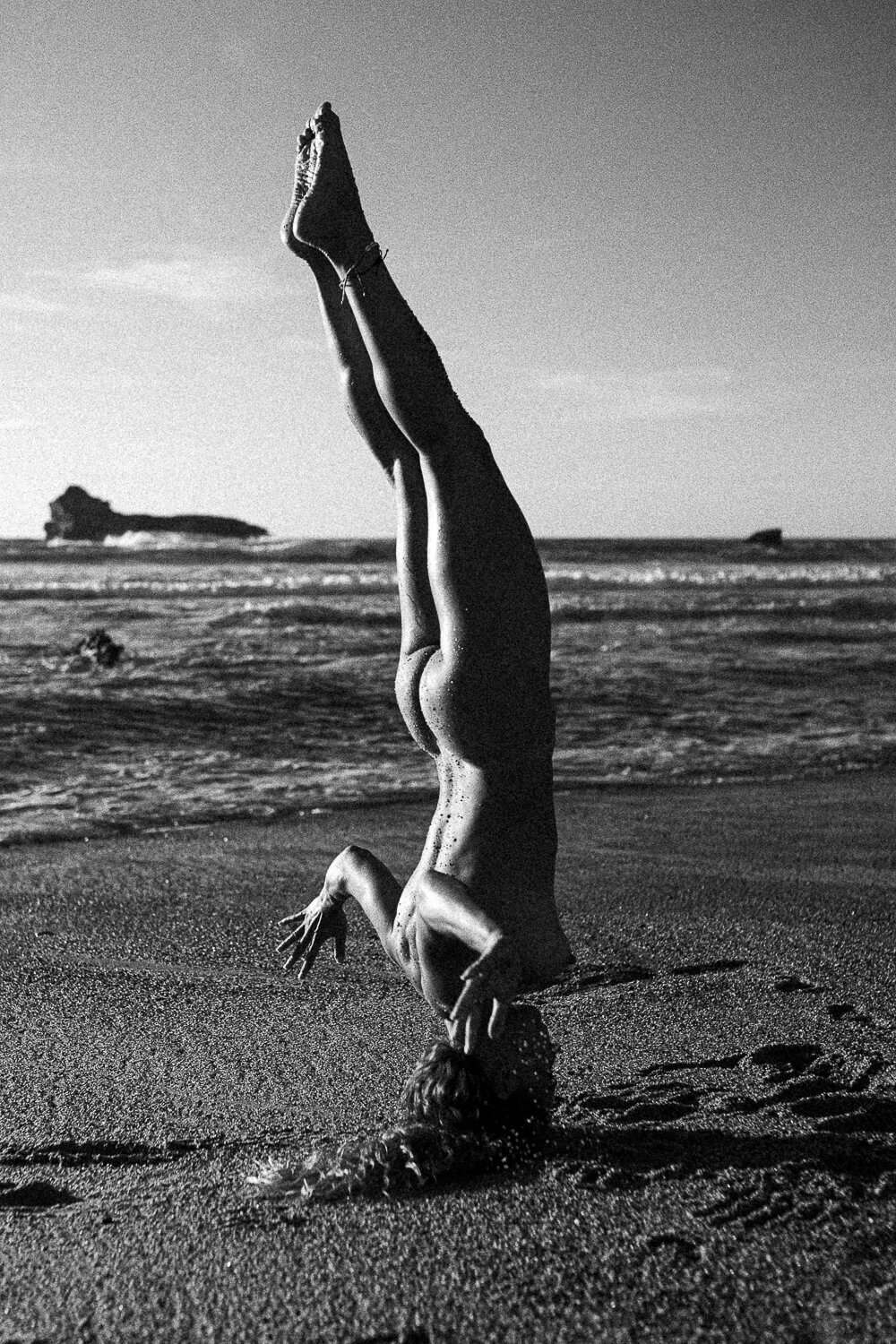 A person performing a handstand on the beach during sunset, with the ocean and a distant rocky formation in the background.