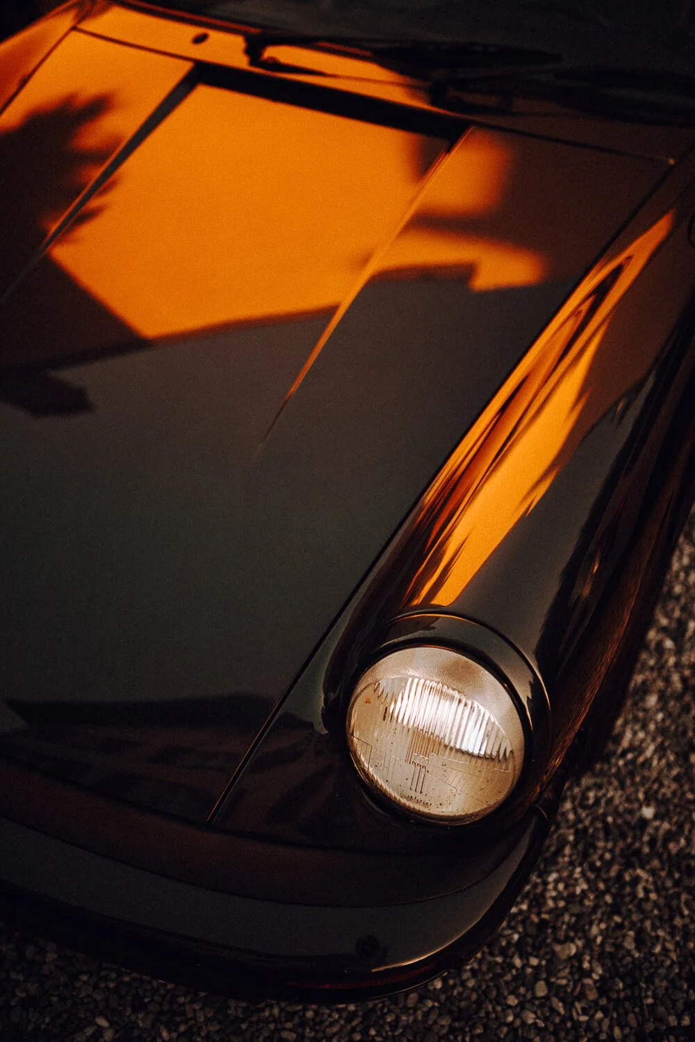 Close-up of the front part of a black sports car with a headlight, reflecting the orange sunset sky.