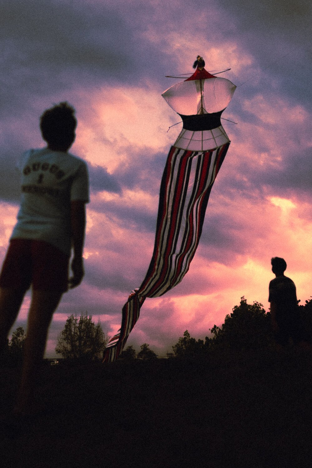 Deux enfants regardant un cerf-volant coloré en forme de personnage dans un ciel au coucher du soleil.