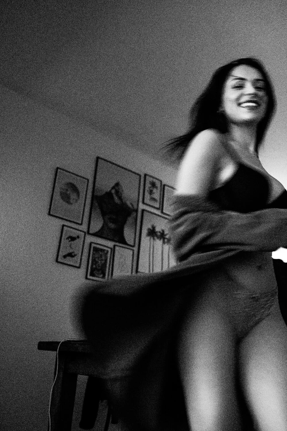 Black and white photo of a smiling woman in underwear standing near a wall with framed pictures and artwork, in a dimly lit room.