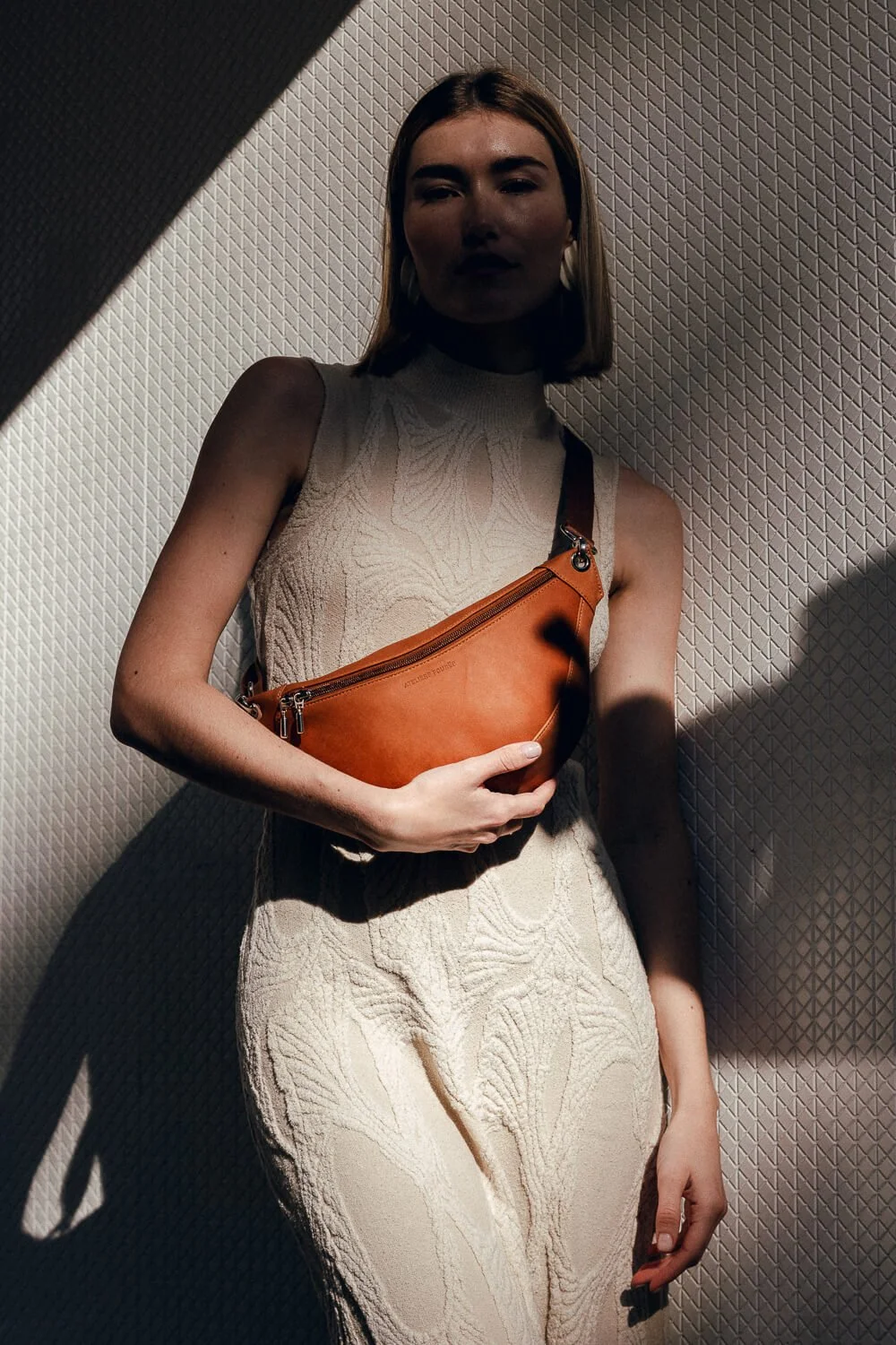 A woman with shoulder-length hair wearing a sleeveless beige dress holding an orange crossbody bag, standing against a textured wall with shadow effects.