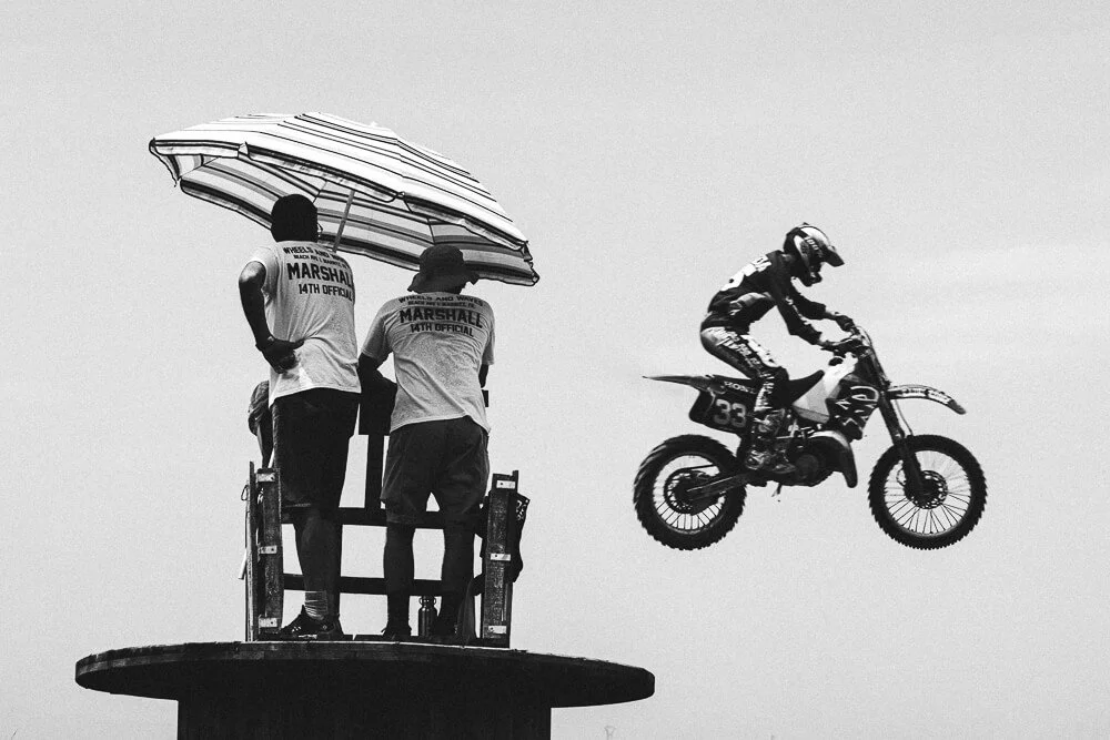 Two officials standing on a platform with an umbrella, observing a motocross rider in mid-air during a jump.