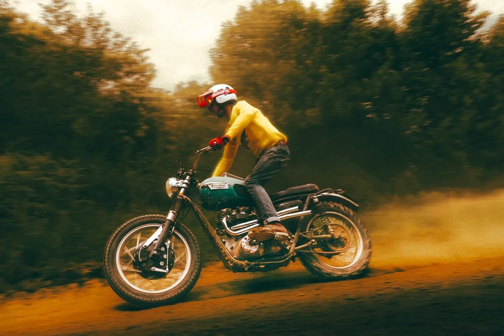 Person riding a vintage motorcycle on a dirt trail, wearing a yellow jacket, helmet, and gloves, with trees in the background and dust kicking up from the tires.