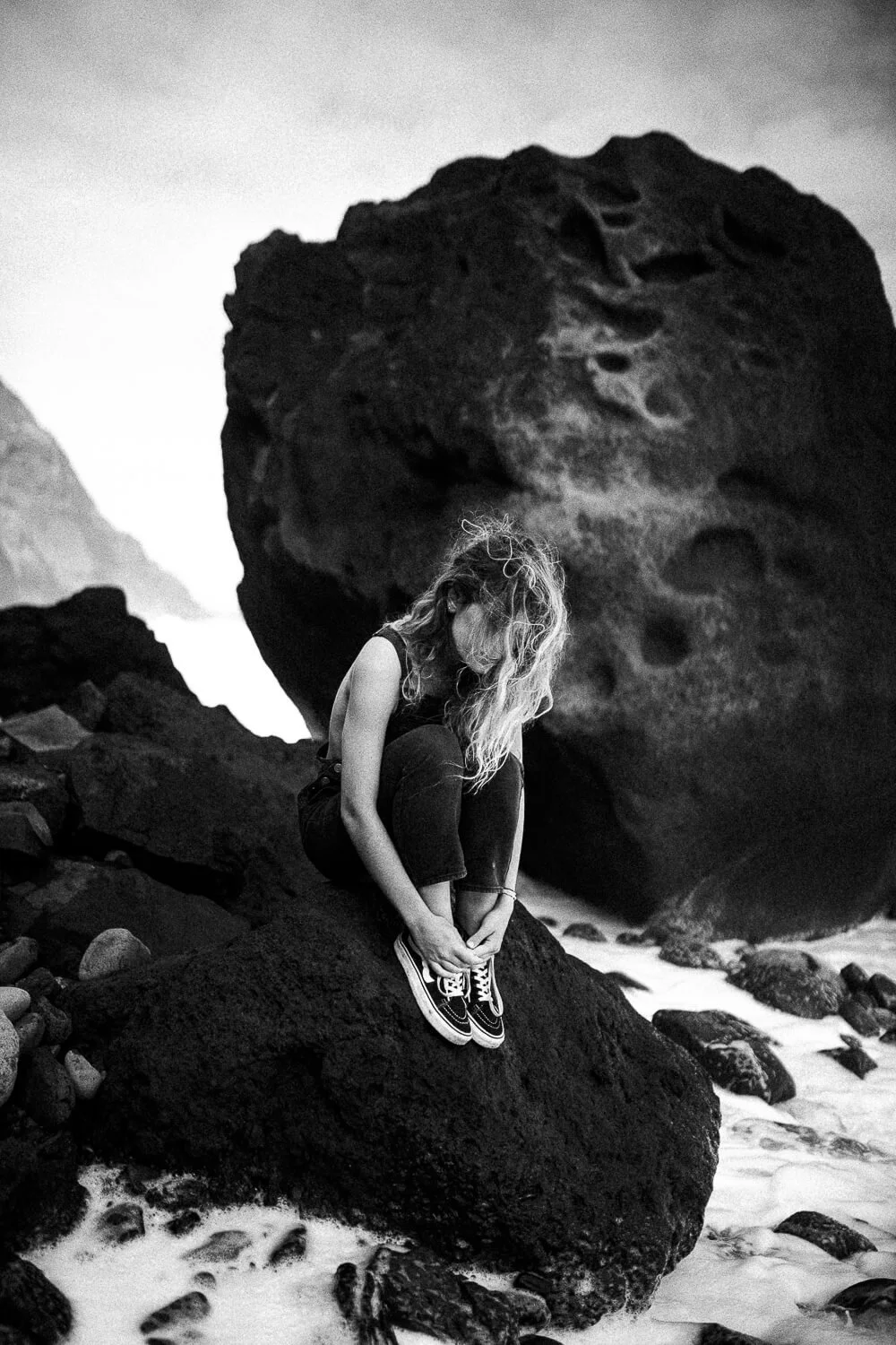 A woman with long, curly hair sitting on a large rock near the ocean, with volcanic rocks and a large, textured boulder behind her, in a black-and-white photo.