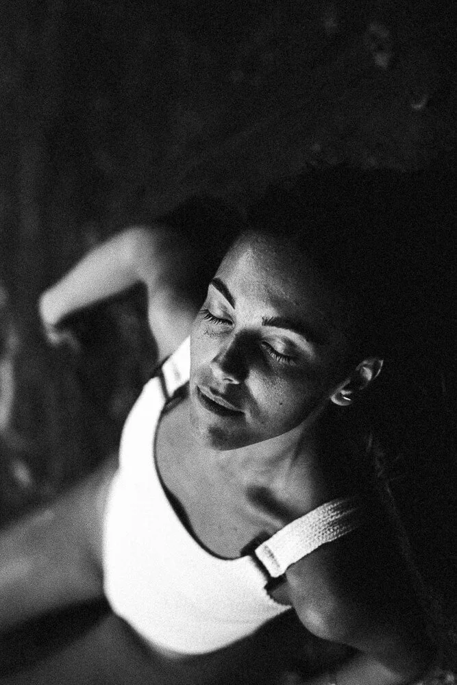 Black and white photo of a young woman with closed eyes, looking down, wearing a sleeveless top, shot from above.