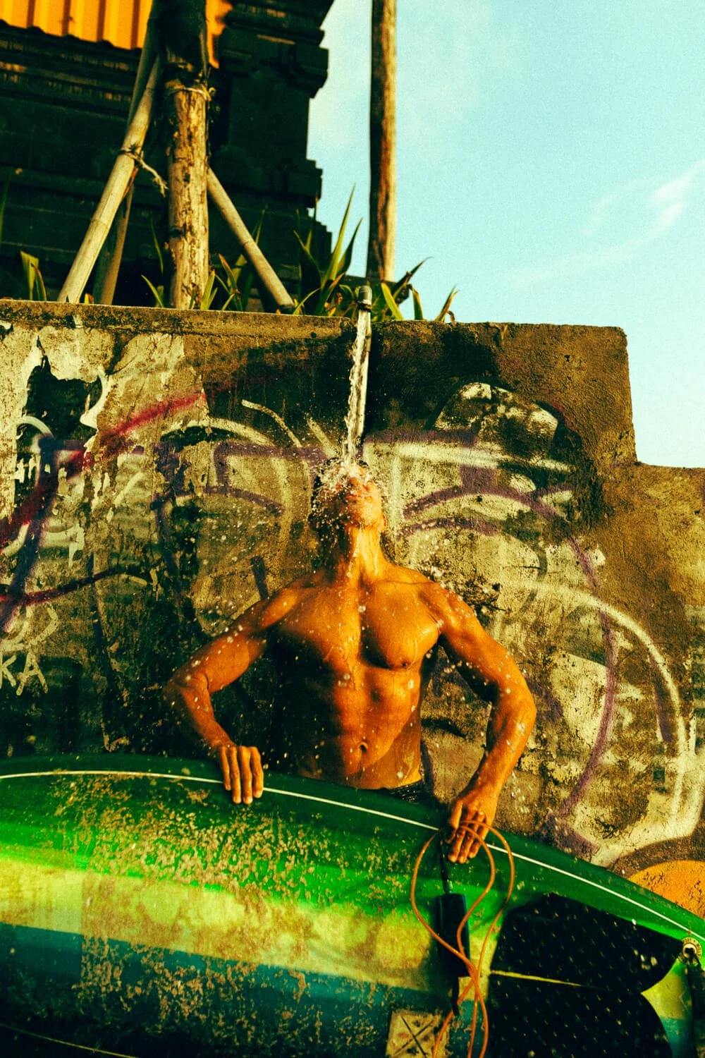 Un mur graffité avec une photo ressemblant à une personnalité célèbre. En premier plan, une personne nue avec la tête sous un jet d'eau, tenant une planche de surf verte. La scène se déroule en plein air sous un ciel bleu.