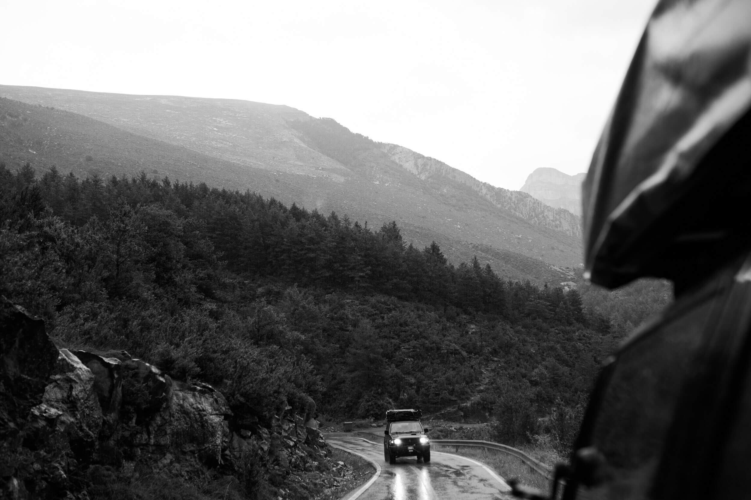 Voiture sur une route de montagne devant une forêt avec des montagnes en arrière-plan, en noir et blanc.