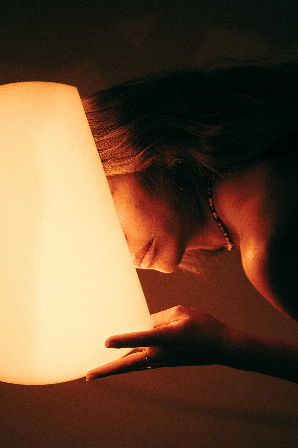 A woman with long hair holding a glowing lampshade close to her face, eyes closed, in a dark room.