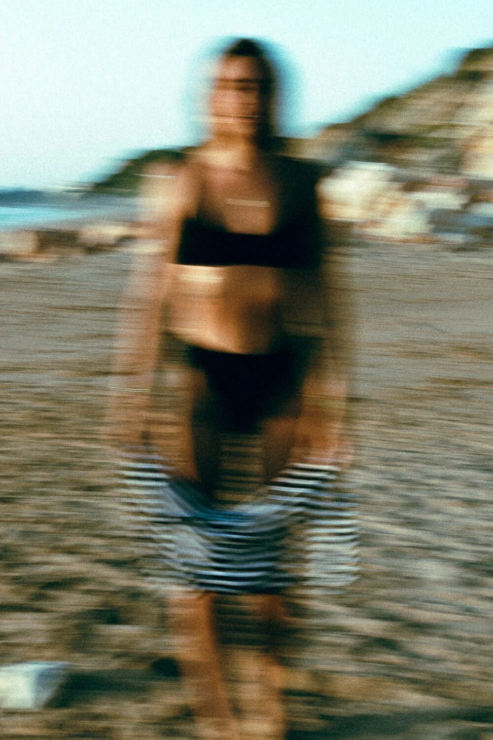 Blurred photo of a woman standing on a beach, wearing a black bikini top, striped shorts, with a tower and rocky hillside in the background.