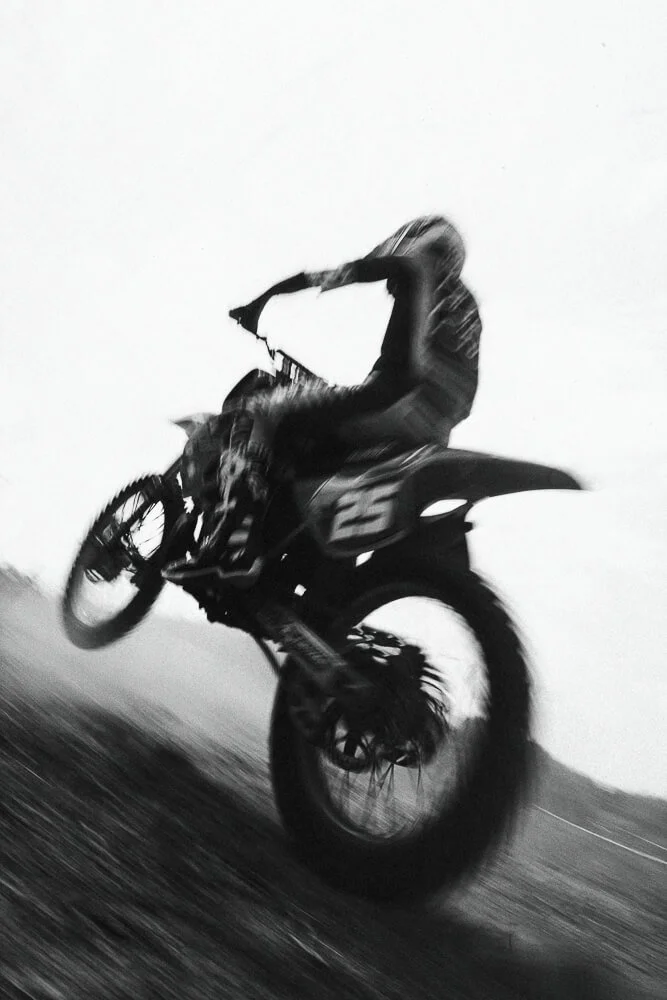 A person riding a dirt bike or motocross motorcycle on a dirt track, captured in black and white with motion blur, showing the rider mid-air during a jump.