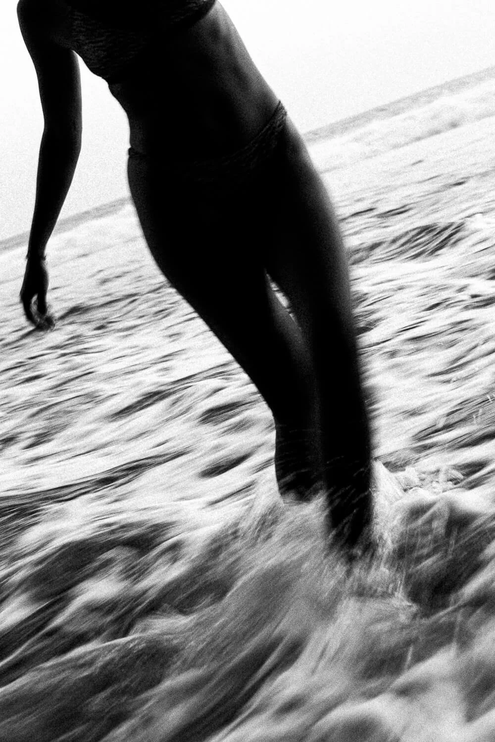 A person standing in the ocean water, shot in black and white, with part of their body and legs visible, as they walk through the waves near the shoreline.