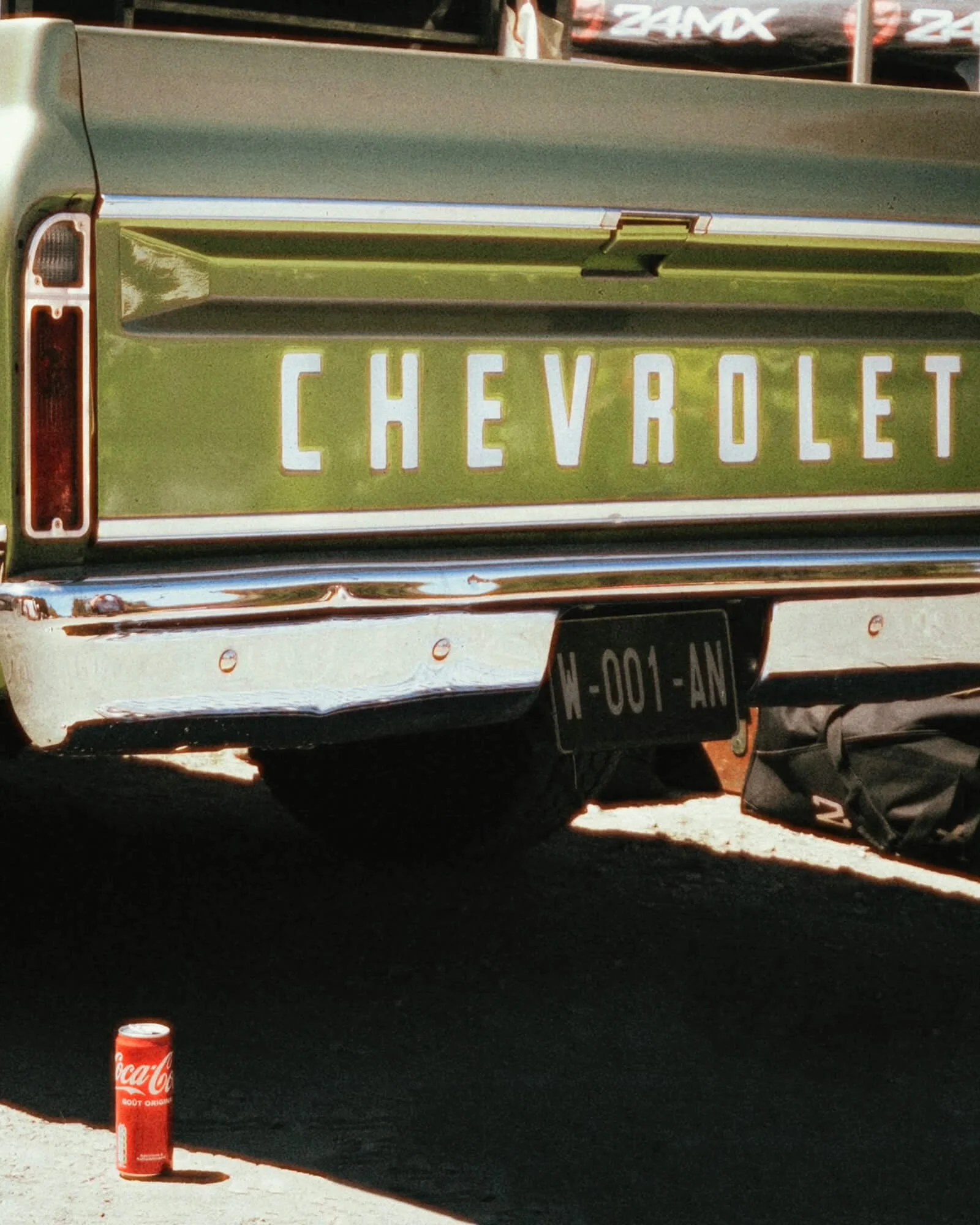 The rear view of a vintage Chevrolet car with a green and silver body, showing the Chevrolet license plate, parked outdoors. A Coca-Cola can is on the ground nearby.