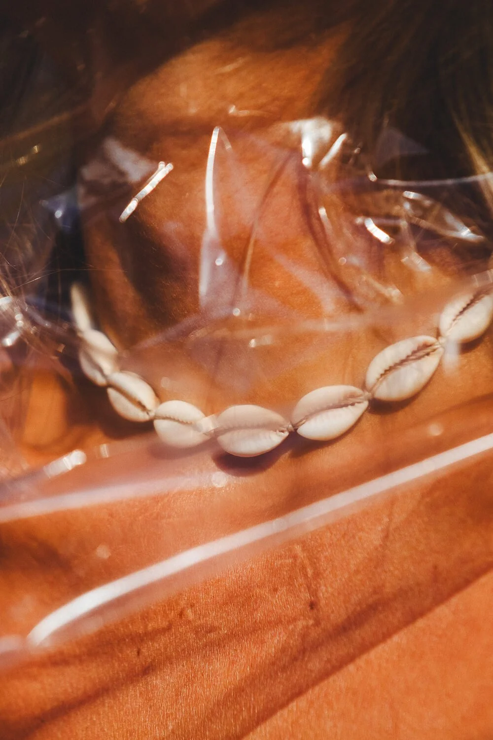 A person wearing a shell necklace and a bra, covered with a sheer orange fabric, under a transparent plastic sheet.