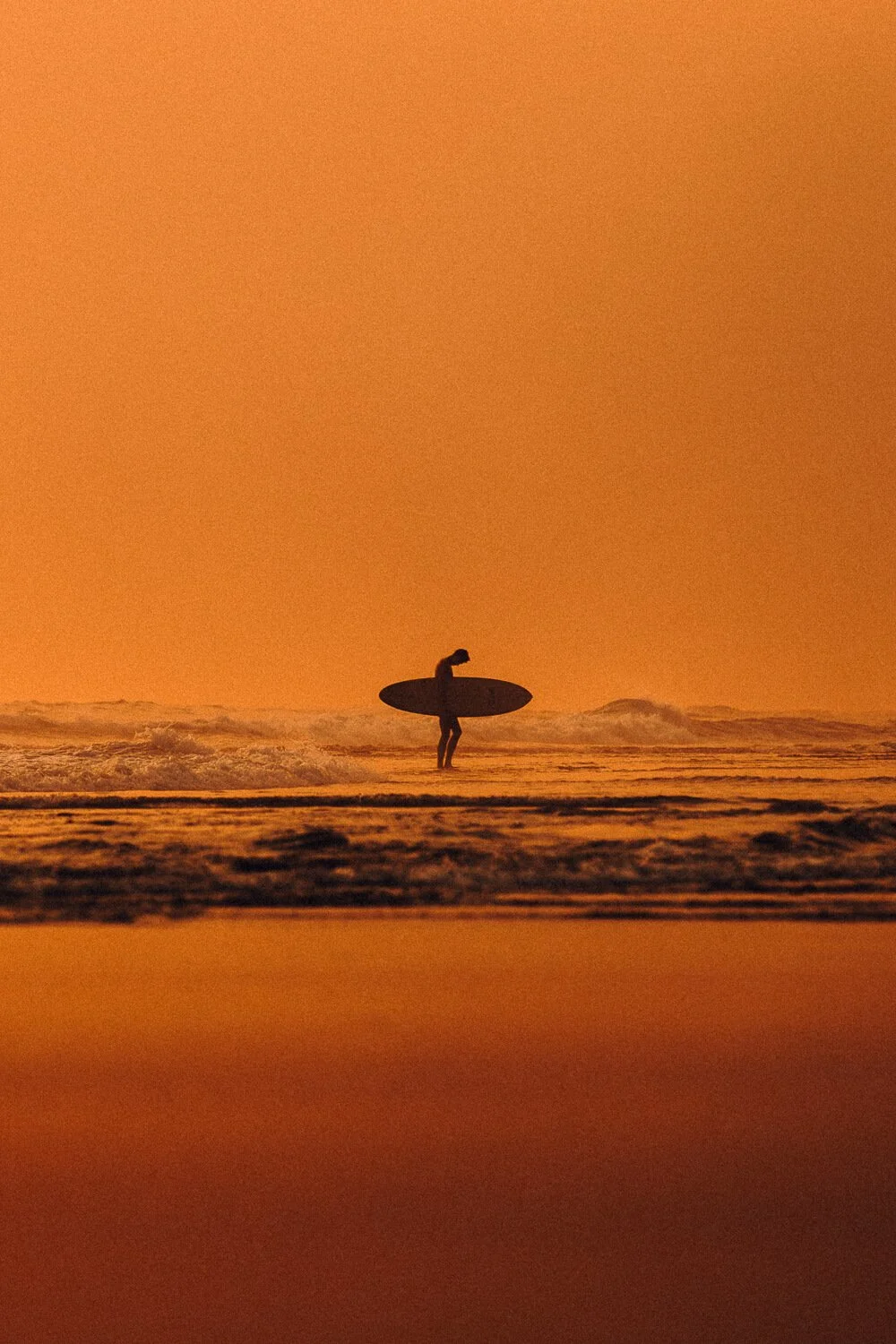 Un surfeur portant une planche de surf se tient dans la mer au coucher du soleil, avec un ciel orangé.