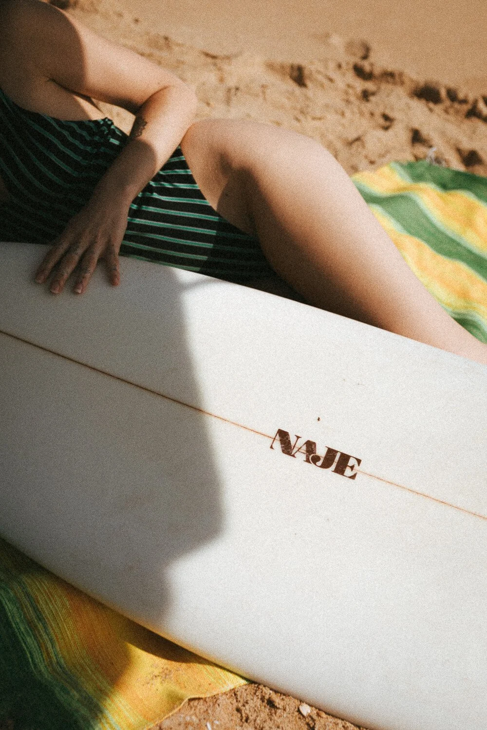 A person laying on the sand at the beach next to a surfboard with the word 'NAE' on it, wearing a striped swimsuit, with a colorful towel nearby.
