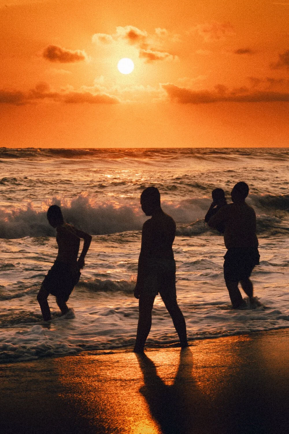 Quatre enfants jouent sur la plage pendant un coucher de soleil, avec le soleil visible dans le ciel orange et des nuages aux alentours.