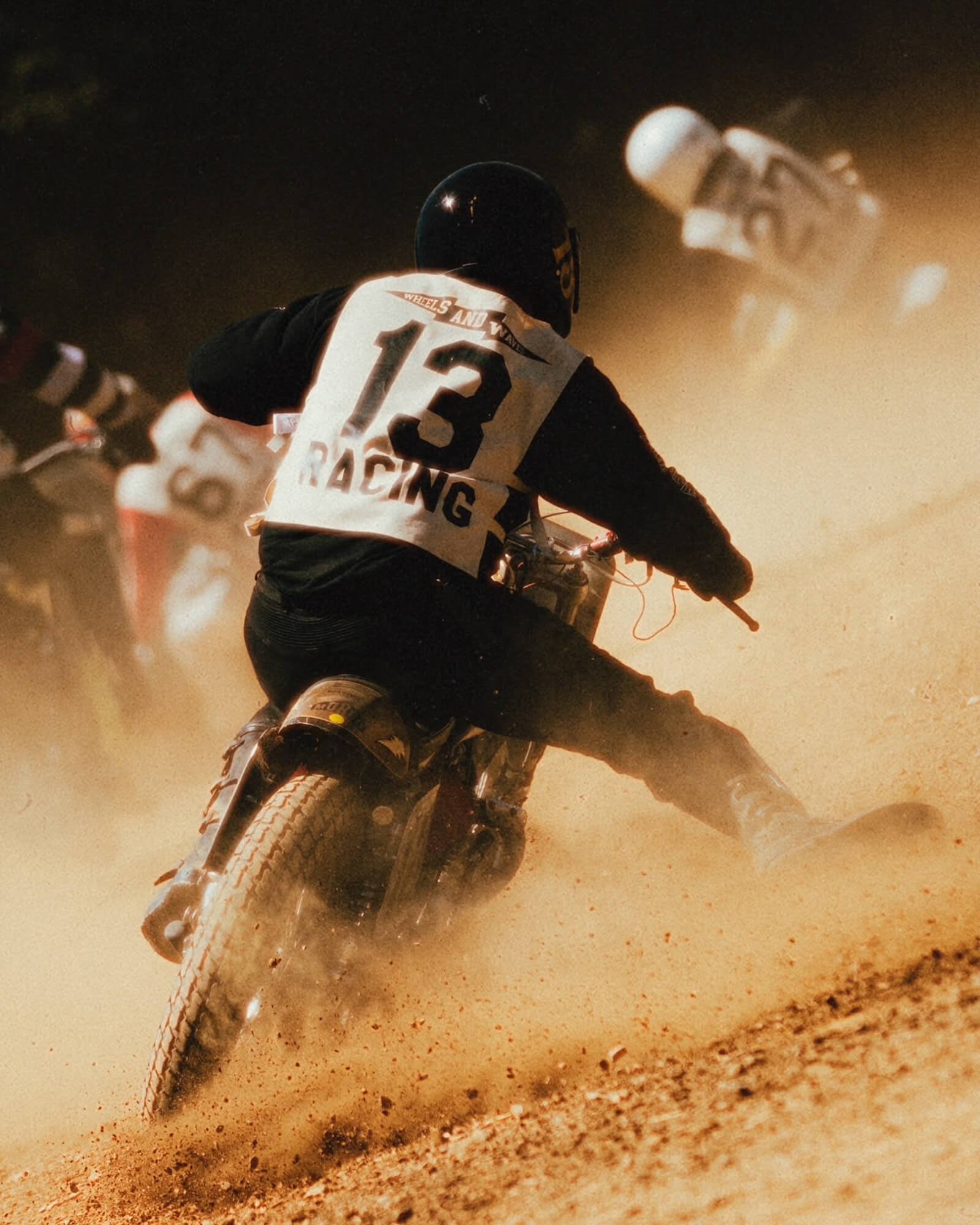 A person riding a dirt bike on a dusty trail, wearing a helmet and racing gear with the number 13 on the back, kicking up dust and dirt behind them.