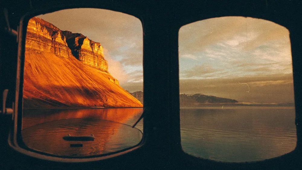 Vue d'une mer calme avec des montagnes et un ciel partiellement nuageux, vue à travers deux fenêtres d'un véhicule ou d'une cabine.