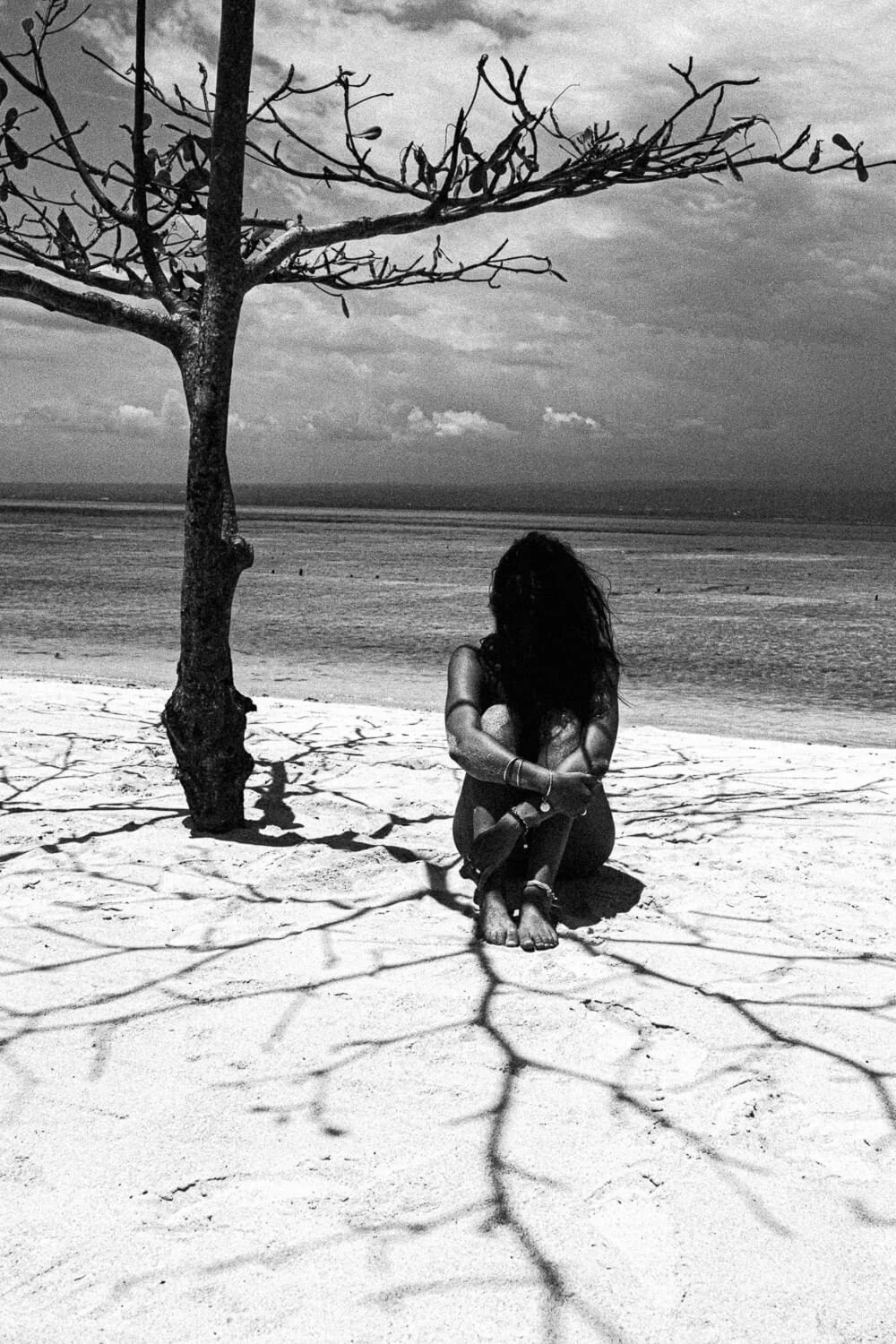 Femme assise sous un arbre dépourvu de feuilles sur une plage, avec des ombres de branches sur le sable, en noir et blanc.