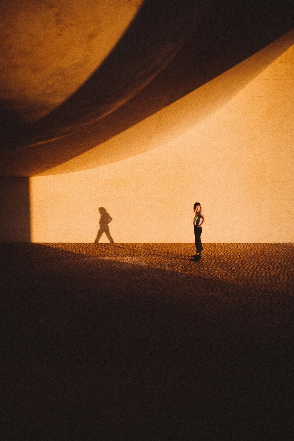 A woman standing on a textured ground with warm lighting creates a shadow of herself on a wall, under a large curved structure.