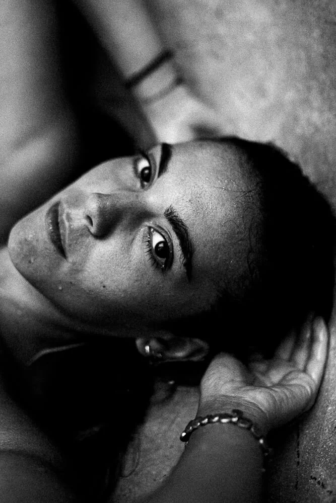 Close-up black and white photo of a woman lying on the floor, with her face turned slightly toward the camera, looking directly at the lens, with her hand resting on her head.