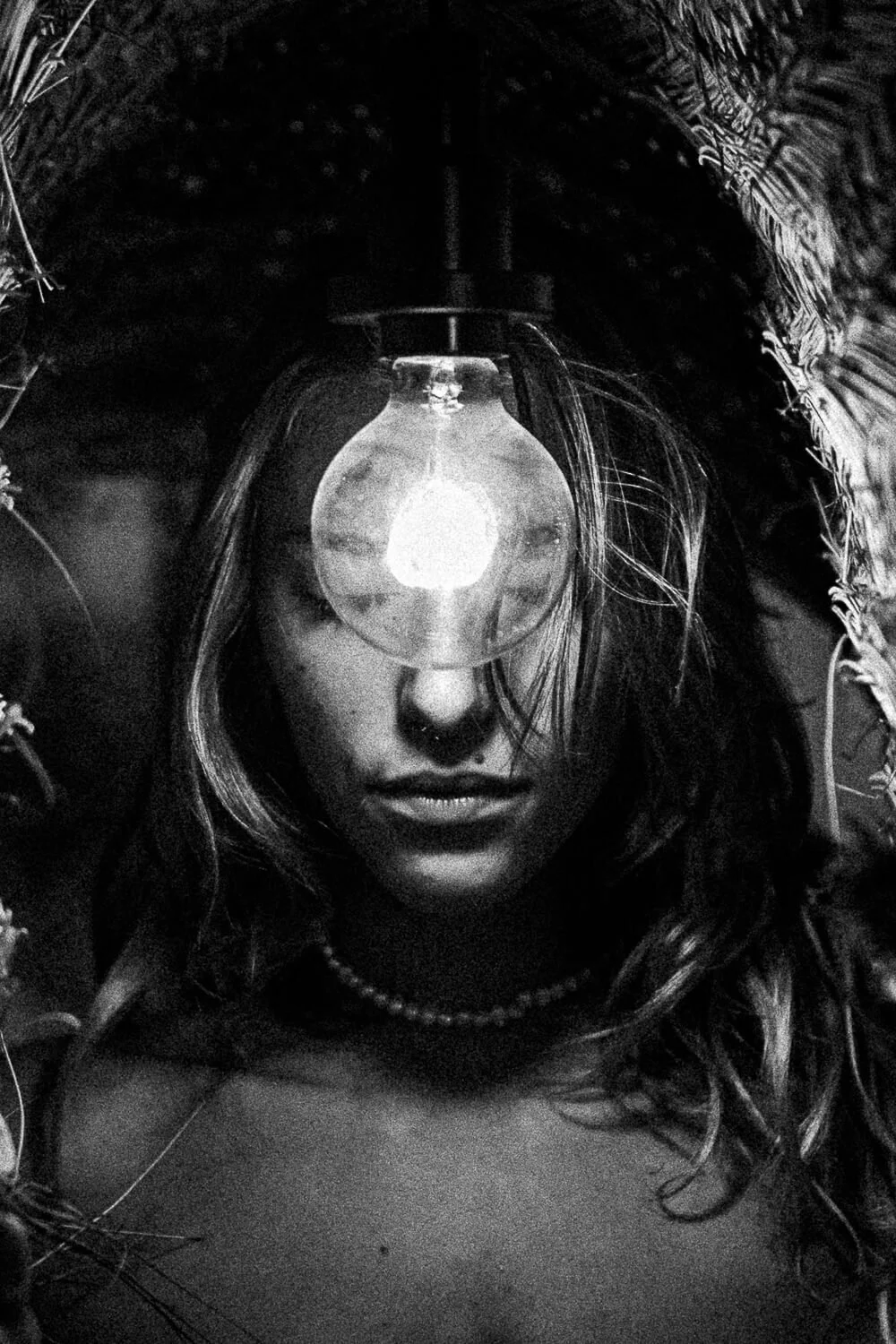 A close-up black and white photo of a woman with long wavy hair, wearing a pearl necklace, with a lit light bulb positioned on her forehead, surrounded by foliage.