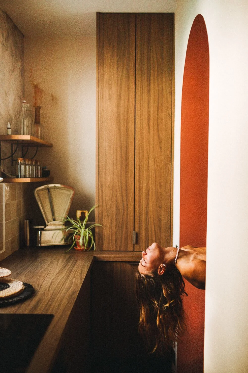 A woman with long wavy hair is leaning back upside down through an opening in a dark wooden cabinet or door frame, in a kitchen setting. The background shows a countertop, a potted plant, and a vintage scale.