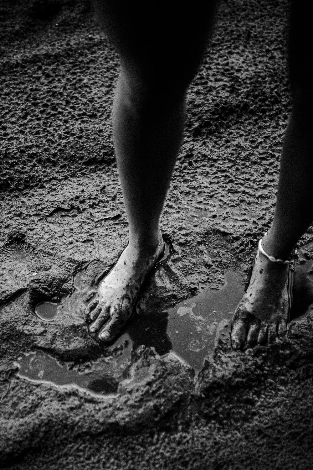Close-up of muddy feet and legs of a person standing on wet, muddy ground, with small puddles of water around.