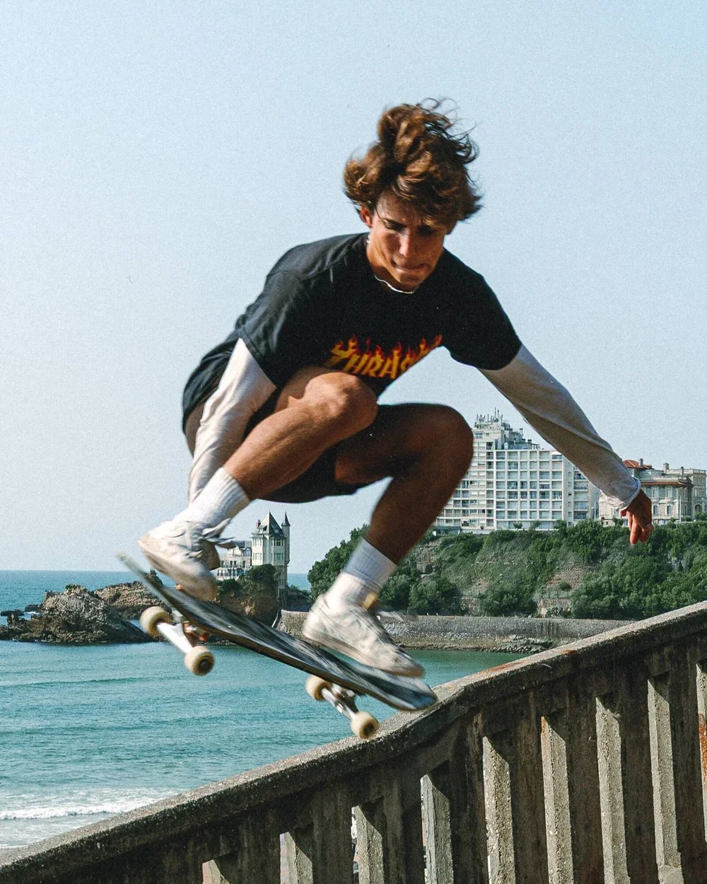 Un jeune homme fait du skateboard sur une rambarde en bord de mer, avec des bâtiments en arrière-plan.