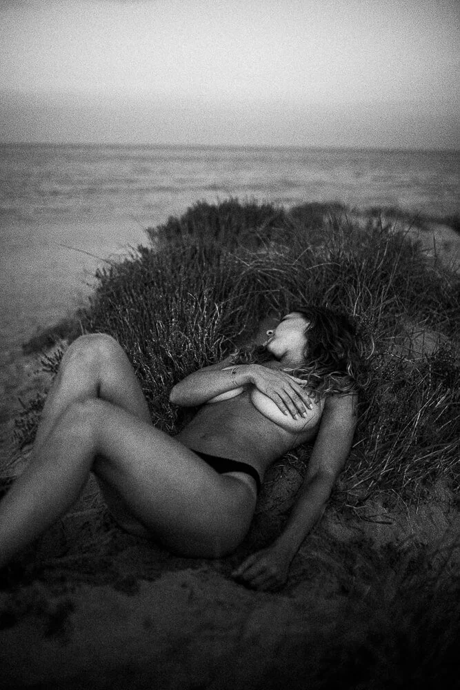 A woman lying on a sandy beach surrounded by tall grass, wearing black underwear, with her arms crossed over her chest and her eyes closed, near the ocean in the background.