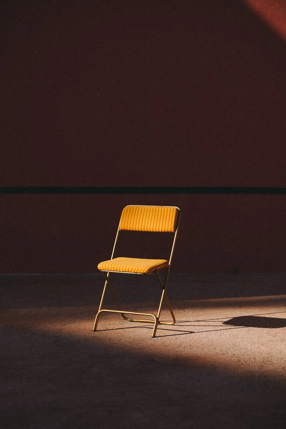 A single vintage patterned orange and yellow striped chair illuminated by a spotlight on a dark stage or room, casting a shadow.