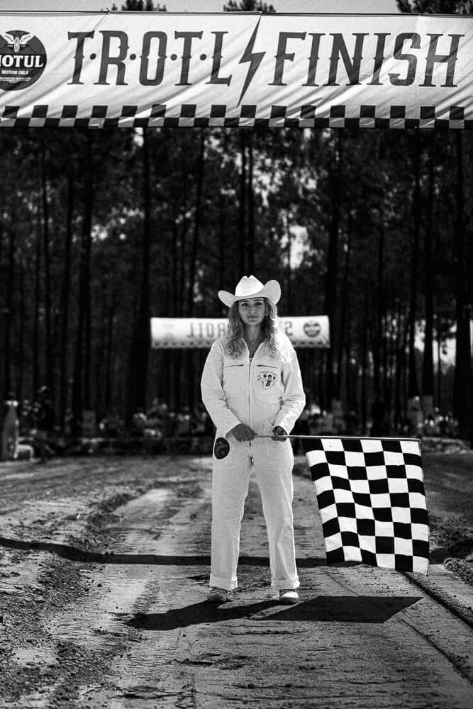 A woman dressed in a white racing jumpsuit and cowboy hat holding a checkered flag, standing on a dirt track under a banner that reads 'TROTL FINISH.'