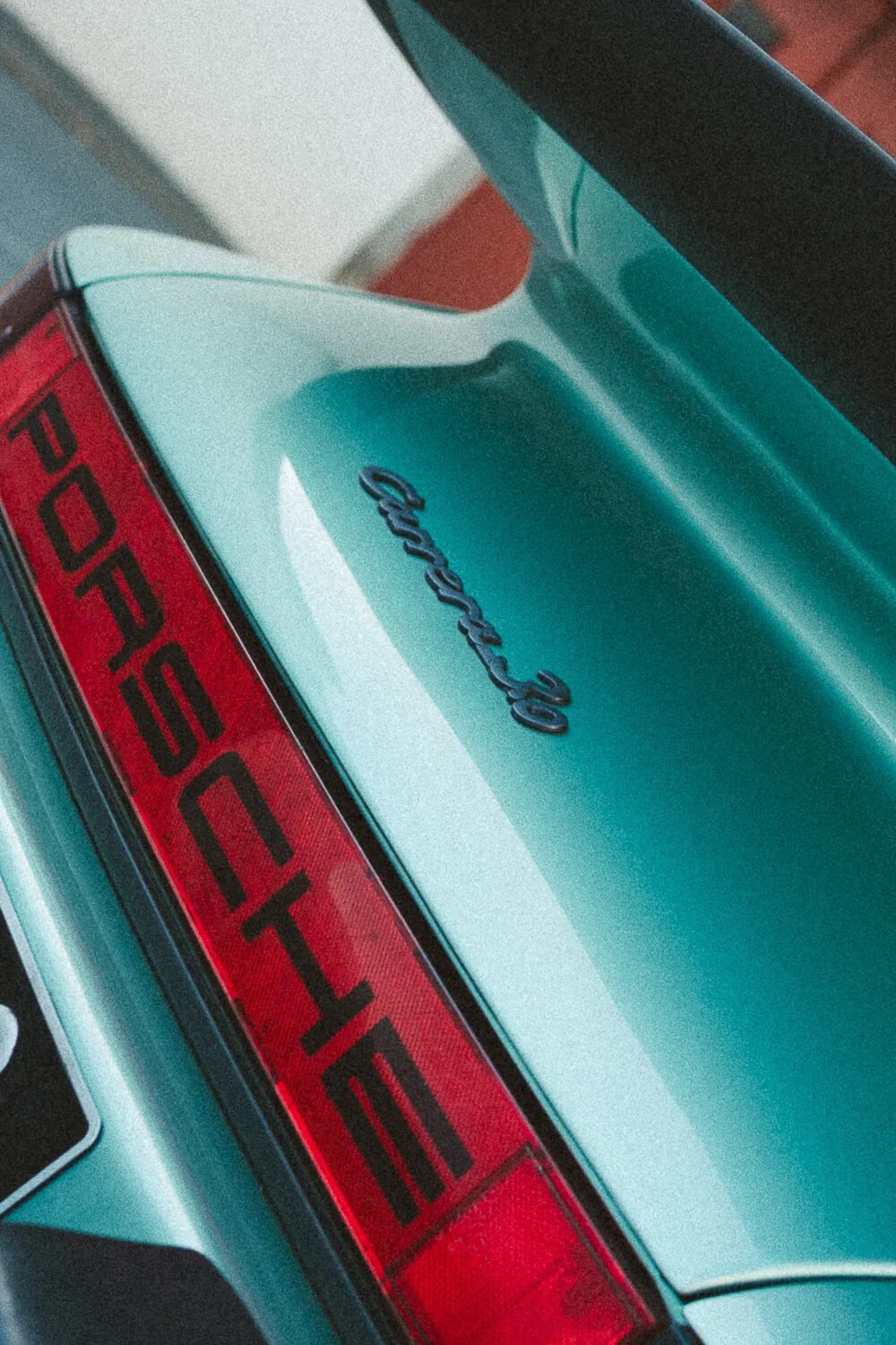 Close-up of the rear end of a teal-colored Porsche 911 Carrera, showing the Carrera badge and Porsche lettering on the red taillight.