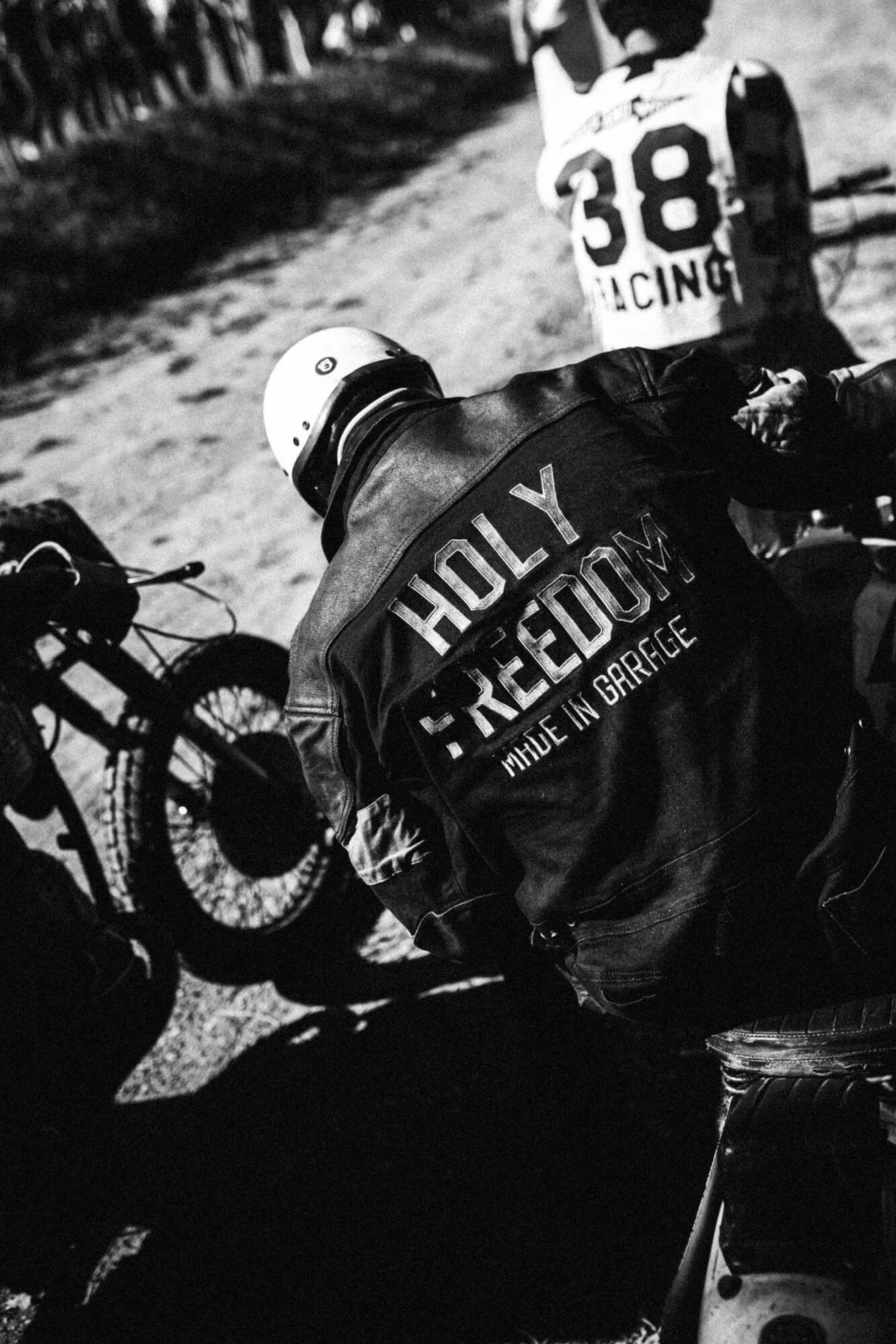 A person wearing a leather jacket with the words 'HOLY FREEDOM MADE IN GARAGE' on the back, riding a motorcycle on a street at night. The person is also wearing a helmet, and another motorcycle is visible nearby.