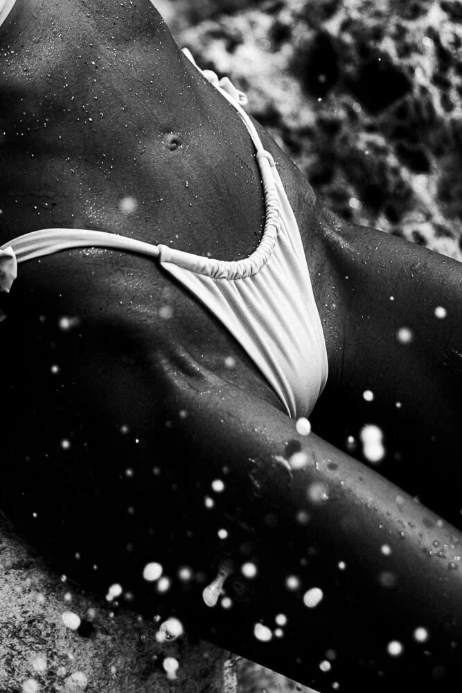 Close-up of a person in a swimsuit, lying on a rocky surface with water splashing around, captured in black and white.