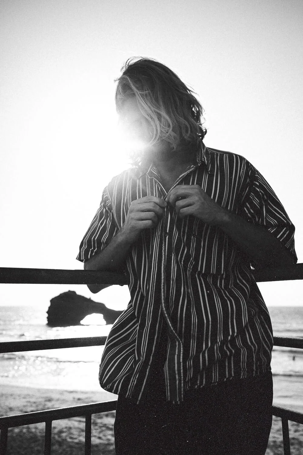 A person with shoulder-length hair stands outdoors near a railing, adjusting their striped shirt, with the sun setting or rising behind them and a large rock formation in the background by the ocean.