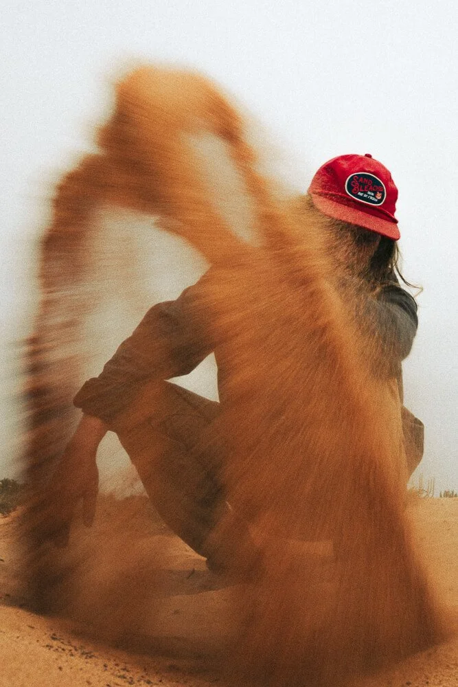 Person sitting on sand with a long-haired dog. The person is wearing a red cap with a logo and has long hair, and the dog is in motion, causing a blurred effect.