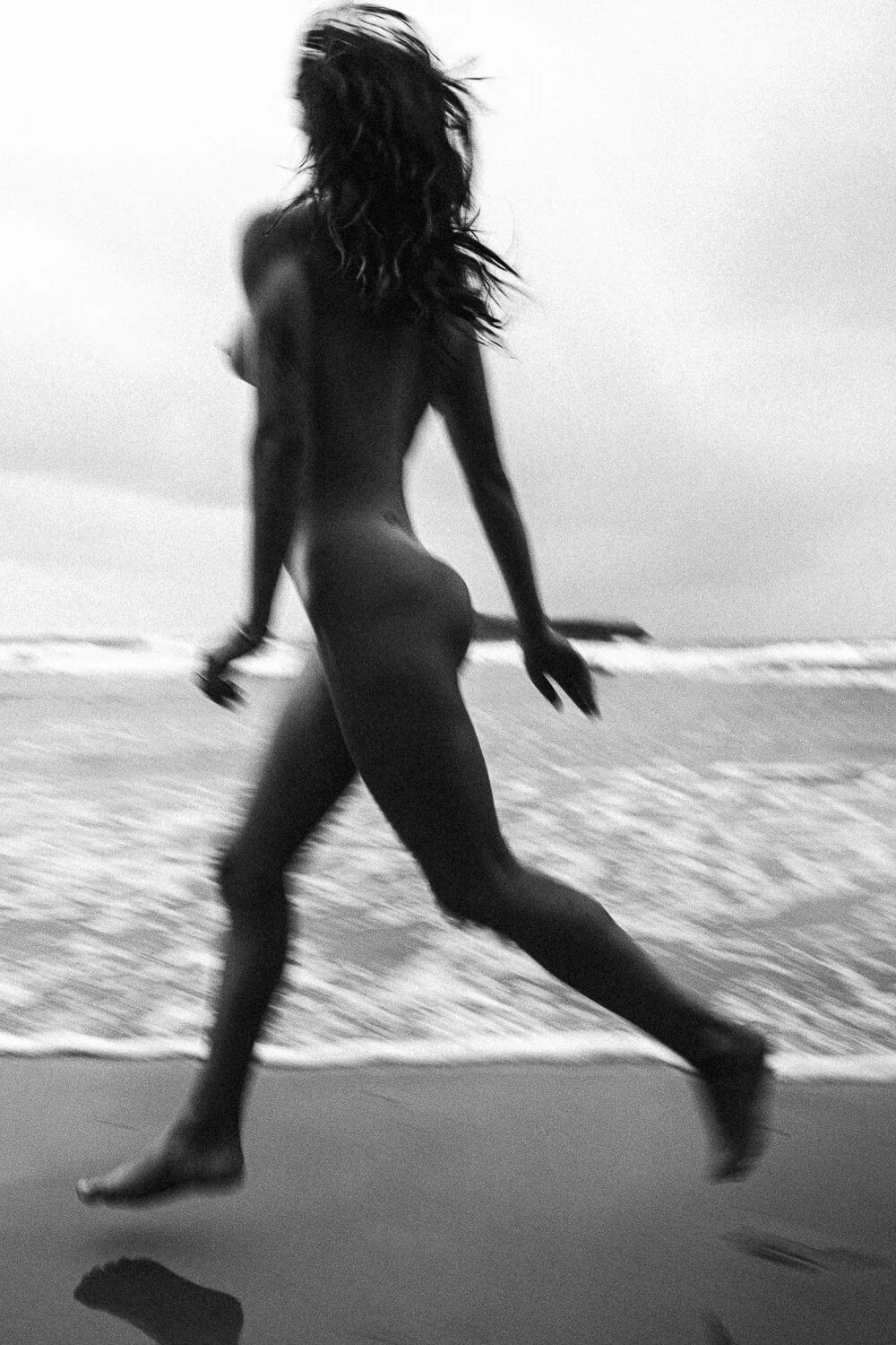 A black and white photograph of a nude woman walking on the beach near the ocean, with her hair flowing in the wind.