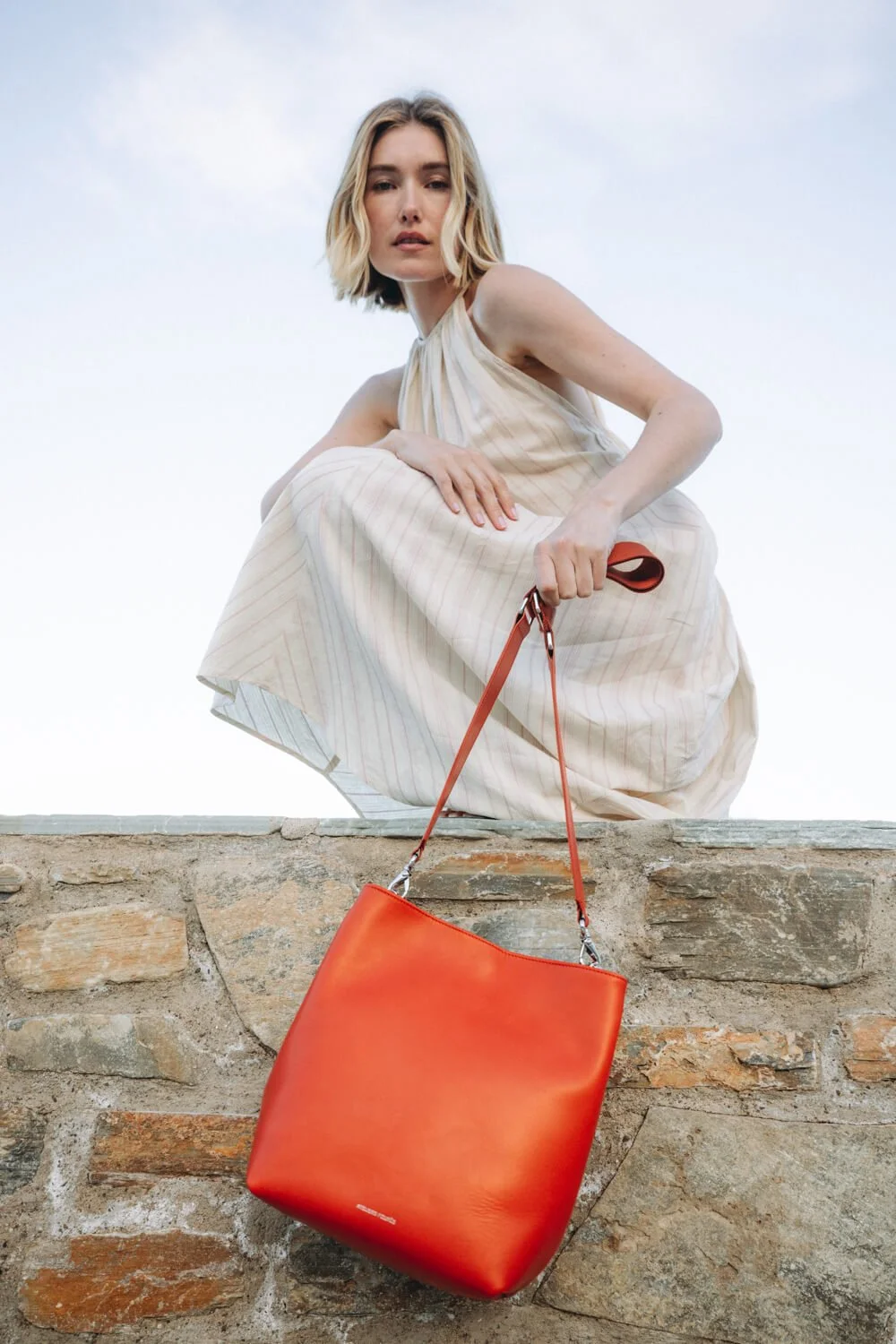 A woman crouching on a stone wall, wearing a beige dress, holding a red leather handbag, against a cloudy sky background.