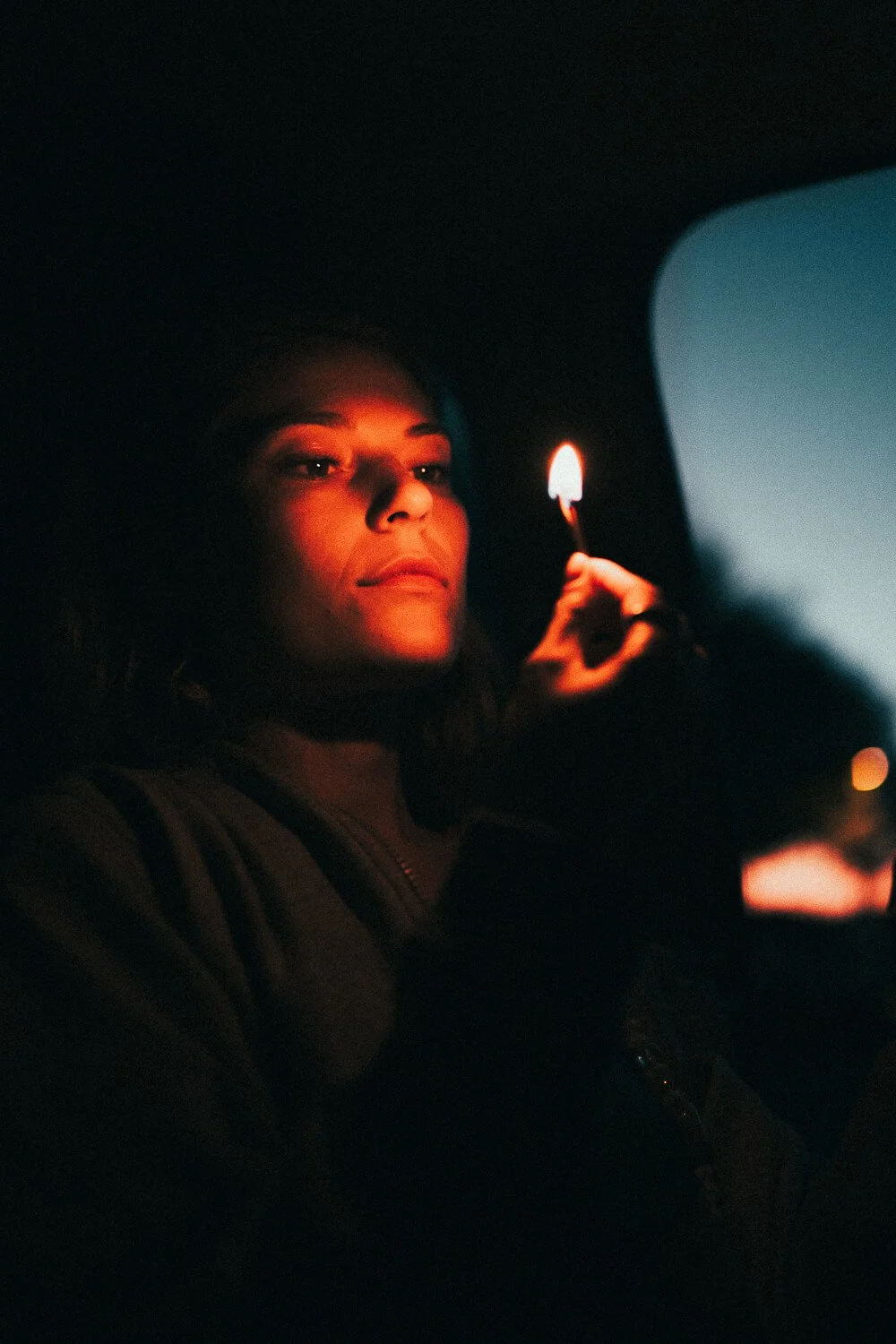 A woman with shoulder-length hair looks out from inside a car at dusk, holding a lit match close to her face, which is illuminated by the match's flame.