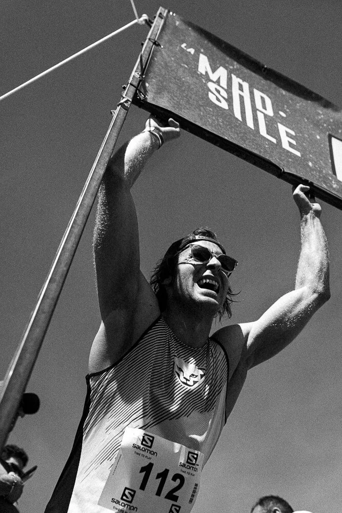 A man wearing sunglasses and a striped tank top, with a race bib number 112, is celebrating victory by holding a large sign overhead at an outdoor event.