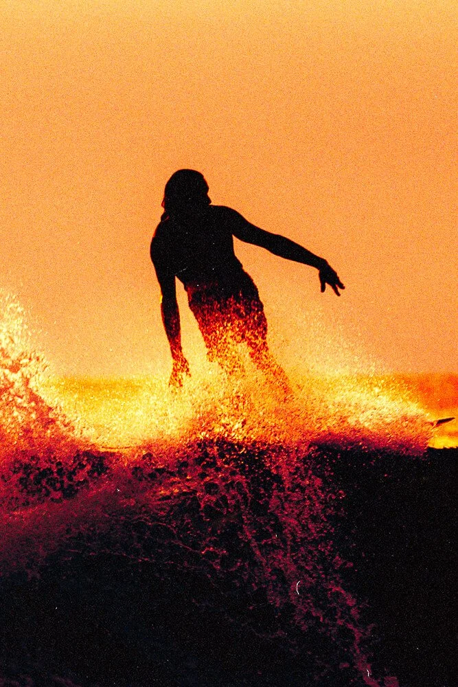 Silhouette of a person surfing during sunset with orange sky and water splashing.
