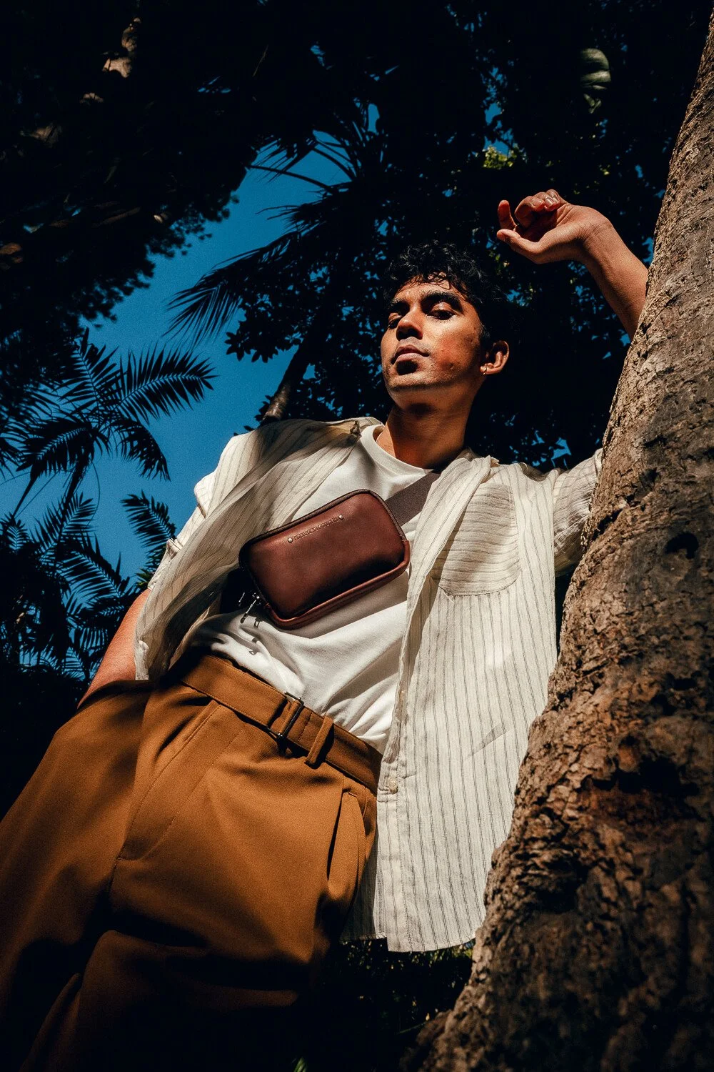 A man leaning against a tree outdoors with tropical foliage and blue sky in the background, wearing a white shirt, beige trousers, and a brown crossbody bag.