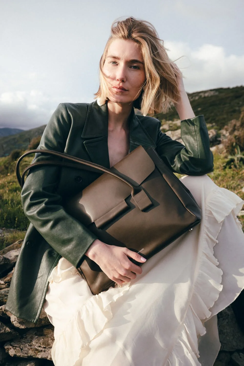 A woman sitting outdoors on rocks with a scenic landscape of hills and sky, holding a large brown handbag, wearing a black leather jacket and a white ruffled skirt.